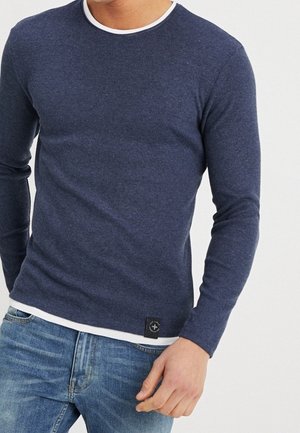 Homme portant un pull ajusté à manches longues bleu marine par-dessus une chemise blanche, associé à un jean bleu clair, sur un fond uni.