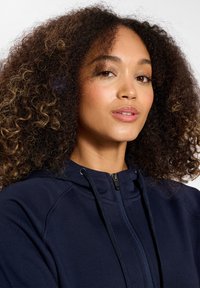 Felpa con zip di colore blu navy scuro, realizzata in un morbido tessuto testurizzato, con un alto colletto e coulisse. Accentuata da una zip frontale e cuciture sottili.