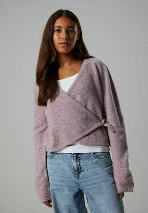 Roze wikkeltrui in een zachte textuur, met lange mouwen en een strikdetail aan de zijkant, gelegd over een witte tanktop en gecombineerd met lichte jeans.