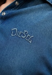 Modrá rifľová košeľa s golierovým výstrihom a kovovými patentkami. Na hrudi má výrazné biele vyšívané logo "DIESEG".