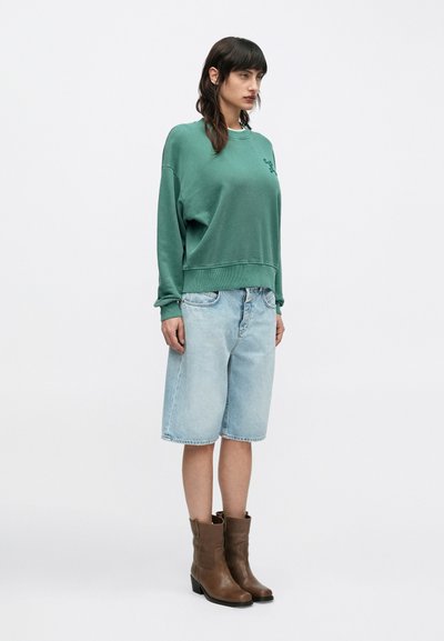 Γυναίκα με πράσινο φούτερ, ανοιχτόχρωμα τζιν σορτς μέχρι το γόνατο και καφέ μπότες μέχρι τη μέση της γάμπας, στέκεται μπροστά σε λευκό, απλό φόντο.