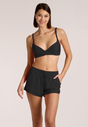 Lächelnde Frau in schwarzem Bikini-Oberteil und schwarzer Badeshorts, die mit einer Hand in der Tasche vor neutralem Hintergrund posiert.