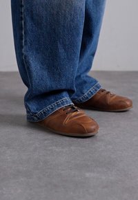 Barna bőr cipő sima textúrával és kerek orral, barna fűzőkkel. Hamvas kék, lazán illeszkedő farmernadrággal viselve.
