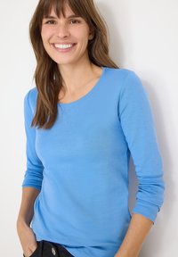 Langspeel, ronde hals trui van lichtblauwe stof, met een gladde textuur en een getailleerd ontwerp. Zwarte broek met zichtbare zakken.