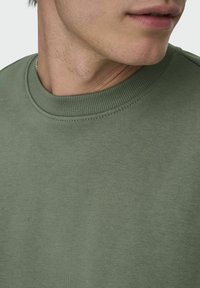 Sweatshirt vert olive en tissu doux, avec un col côtelé et une conception sans couture aux épaules. Gros plan sur le col et les détails de texture.