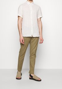 Lněná košile s krátkým rukávem v světlé béžové barvě, spárovaná s světlými hnědými kalhotami na míru a tanovými klouzavými sandály s černými akcenty.