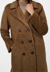 Brun dubbelknäppt kappa i texturerat tyg med svarta knappar, spetsig krage och sidofickor. Mjuk och varm framtoning.