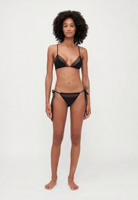 Juodas bikinio komplektas su trikampiu viršumi ir plonais raišteliais, bei šoninėmis raištelėmis surišamais apatinių kelnaitėmis, abiejose dalyse dekoruotomis nėrinių detalėmis.