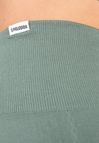 Olivově zelená ribovaná látka s hladkým povrchem a přišitým štítkem s nápisem "SMILODOX" na výstřihu. Design obsahuje horizontální ribbing.