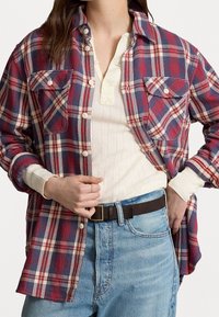 Frau trägt ein rot-blau-weiß kariertes Hemd über einem cremefarbenen Henley-Oberteil, kombiniert mit hellblauen Jeans und einem dunkelbraunen Gürtel.