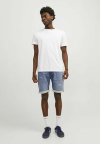Camiseta blanca de cuello redondo combinada con pantalones cortos de mezclilla azul claro con parches desgastados y bajos enrollados. Lleva puestas zapatillas deportivas azul oscuro.