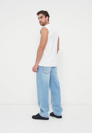 Man wearing sleeveless white shirt, light blue wide-leg jeans, and black sneakers, standing and looking back over his shoulder.