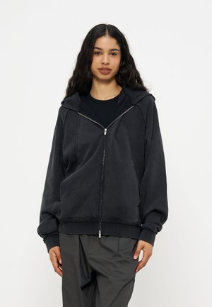 Ung kvinde med langt mørkt hår iført en sort hættetrøje med lynlås over en sort skjorte og mørkegrå bukser mod en ensfarvet hvid baggrund.