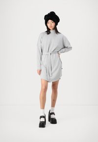 Vestido cinza de sweatshirt com mangas longas, cintura cingida e bolsos laterais. A modelo usa um chapéu de pele preto e sapatos de plataforma pretos chunky.