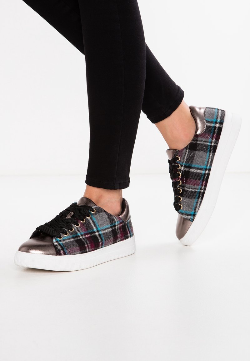 Zapatillas de cuadro con parte superior de tela gris, rayas azules y burdeos, puntera de bronce metálico y cordones negros sobre una suela blanca.