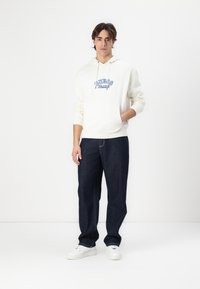 Sudadera blanca con el logo azul de "Guess Jeans", combinada con vaqueros azul oscuro y zapatillas blancas. El modelo se encuentra de pie frente a un fondo liso.