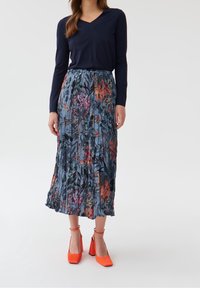 Top blu navy a maniche lunghe, gonna maxi plissettata floreale in tonalità di blu, rosa e verde, abbinata a sandali arancioni a punta quadrata.