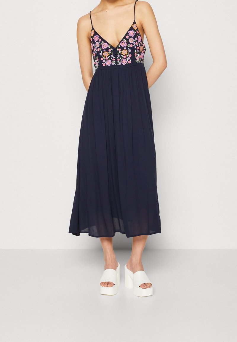 Navyblaues Maxikleid mit floral besticktem Oberteil, V-Ausschnitt und verstellbaren Spaghetti-Trägern. Getragen mit weißen Plateausandalen.