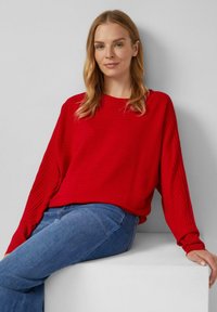 Rotgestreifter, übergroßer Pullover mit langen Ärmeln, Rundhalsausschnitt und lockerer Passform, kombiniert mit blauen Jeans, vor einem neutralen Hintergrund.
