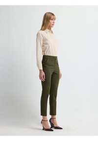Beige overhemd met lange mouwen en knopen, olijfgroene broek met hoge taille, en donkere hakken met banden. Effen achtergrond.