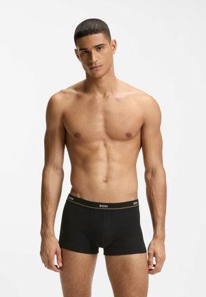 Junger Mann mit kurzen Haaren, trägt schwarze BOSS Boxershorts, steht vor einem schlichten weißen Hintergrund.