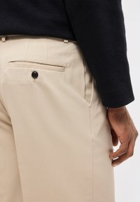 Vue arrière d'une personne portant un pantalon beige avec une poche arrière boutonnée et un haut noir à manches longues, la main reposant près de la couture latérale.