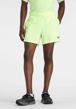 Männliches Modell trägt neongelbe Sportsshorts und schwarze Laufschuhe vor weißem Hintergrund.