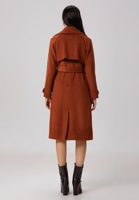 Manteau trench en laine marron avec une taille ceinturée et un design à double boutonnage. Les détails notables comprennent une fente dans le dos et un large col.