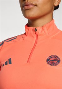 Pink quarter-zip sports top with gray stripes. Features FC Bayern München logo, smooth texture, and close-fitting design.