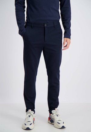Navy-blaue Baumwollmischungshosen mit Slim-Fit, ausgestattet mit einem Knopfverschluss und seitlichen Taschen, kombiniert mit weißen Sneakers mit roten und blauen Akzenten.