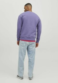 Sudadera morada con cuello redondo, puños y dobladillo de canalé. Usada sobre una capa roja, combinada con jeans de mezclilla azul claro y zapatillas negras.