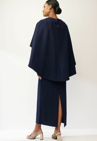 Marinblå cape-klänning med en nyckelhål i ryggen, som har en lång kjol med en sidoklyfta och blanka guldblockklackar. Slät textur och strukturerad design.
