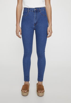 Femme portant un jean skinny bleu taille haute et des sandales à plateformes marron, debout devant un fond blanc uni.
