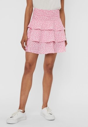 Femme portant une mini-jupe rose à volants avec des motifs floraux blancs et des baskets blanches, debout devant un fond clair uni.