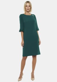 Turkusowa sukienka do kolan z trzyczwartowymi rękawami, falbaniastymi mankietami i dwiema ozdobnymi guzikami na ramieniu. Gładka faktura, prosta sylwetka.