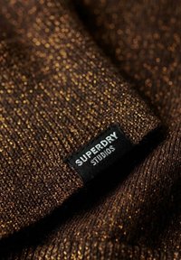 Tessuto marrone con texture e fili metallici dorati; presenta un'etichetta nera con testo bianco che recita "SUPERDRY STUDIOS."