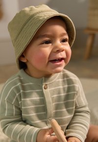 Camicia a maniche lunghe con righe verde chiaro e abbottonatura, abbinata a un cappello a secchiello verde morbido, con un giocattolo di legno in mano.