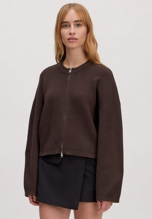 Bruine geribbelde trui met een ritssluiting aan de voorkant, ronde halslijn en oversized mouwen, gecombineerd met een zwarte minirok.