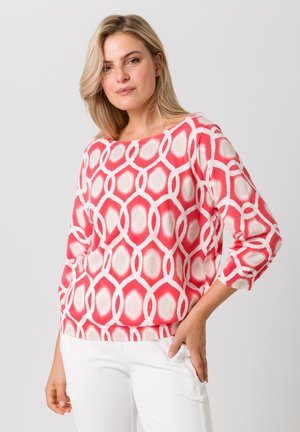 Pull léger rose avec un motif géométrique en blanc et beige clair. Il possède un décolleté large et des manches mi-longues.