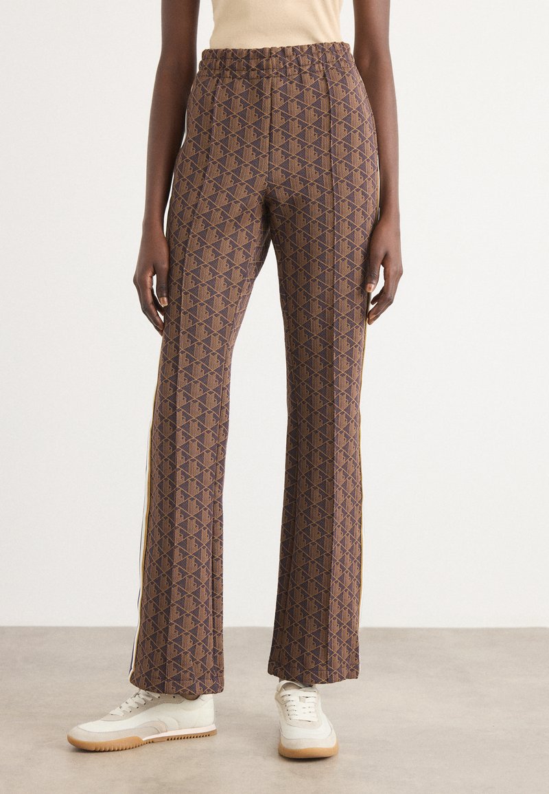 Pantaloni marroni con un motivo geometrico, vita elastica e strisce laterali. Accoppiati con sneakers bianche su un pavimento neutro.