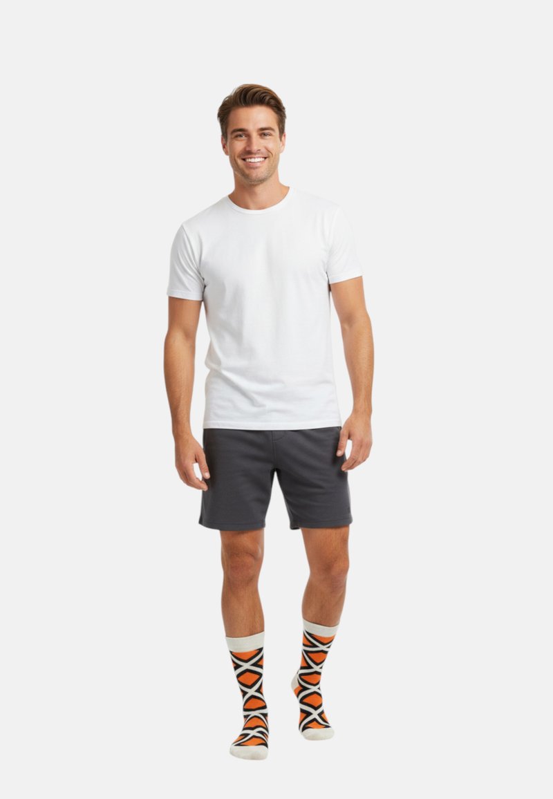 Smilende mand i hvid t-shirt, mørkegrå shorts og orange mønstrede sokker, som står mod en ensfarvet hvid baggrund.