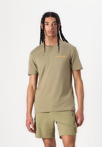 Alyvuogių žalios spalvos medvilnės marškinėliai su apvaliu kaklu ir Quiksilver logotipu. Derinami su atitinkamais cargo šortais, abu turintys lygią tekstūrą.