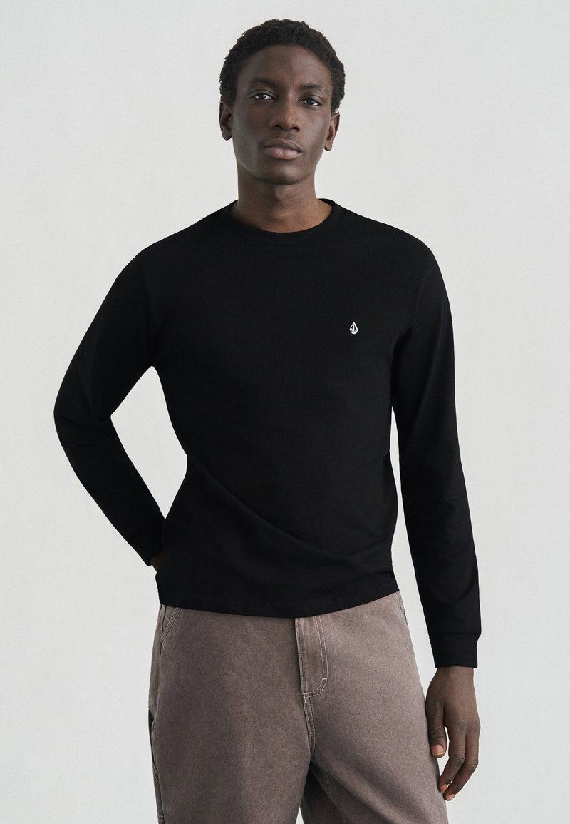 Μαύρο πουκάμισο με μακριά μανίκια και στρογγυλή λαιμόκοψη, με μικρό λευκό λογότυπο στο στήθος. Φορεμένο με ανοιχτό καφέ παντελόνι. Λείο ύφασμα.