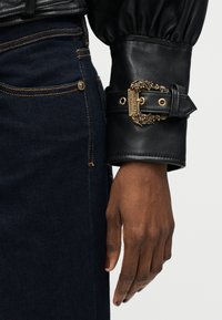 Χέρι με σκούρο δέρμα που φοράει μαύρο δερμάτινο μανίκι με περίτεχνη χρυσή αγκράφα Versace, δίπλα σε σκούρες μπλε τζιν-παντελόνι με ορατές ραφές.