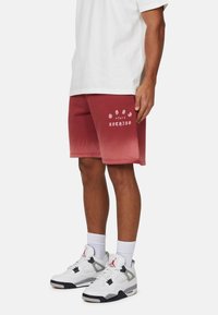 Pantaloni corti con gradiente rosso e testo "bad for nothing" in bianco, abbinati a sneaker bianche con accenti rossi e neri.