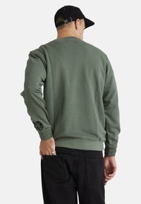 Green crewneck sweatshirt made of soft fabric; features a black graphic detail on the left sleeve. Paired with dark jeans.