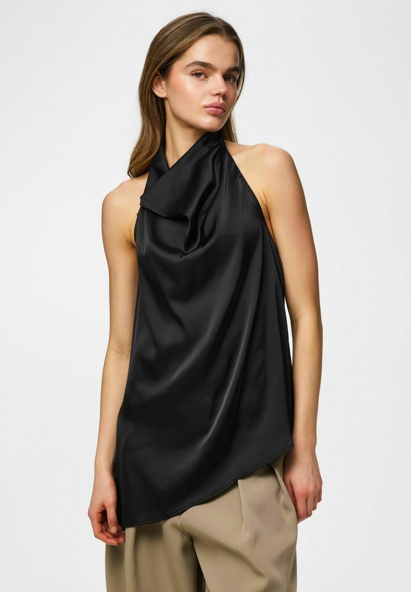 Femme portant un top dos nu en satin noir et un pantalon beige, posant devant un fond blanc uni.