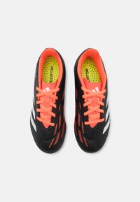 Zapatos de fútbol sintéticos negros con cordones naranjas, con un upper texturizado y rayas blancas. La plantilla es de un amarillo brillante con perforaciones.
