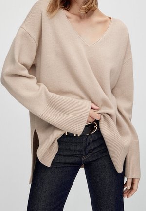 Maglione beige a maglia con scollatura a V, polsini a coste ampie e spacchi laterali. Indossato su jeans scuri con una cintura nera con accenti metallici.