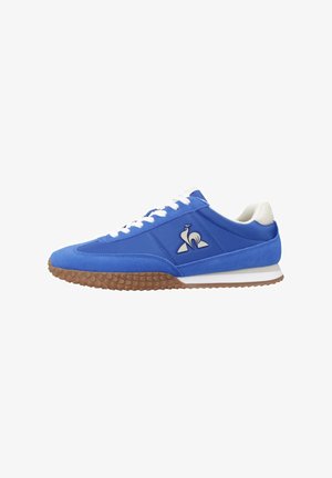 Chaussure de sport bleue en suede et tissu, avec une semelle et des lacets blancs, un logo sur le côté et une semelle extérieure en caoutchouc brun texturé.